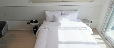 A neatly made bed with white linens, a bedside table with a lamp, and a window with blinds.