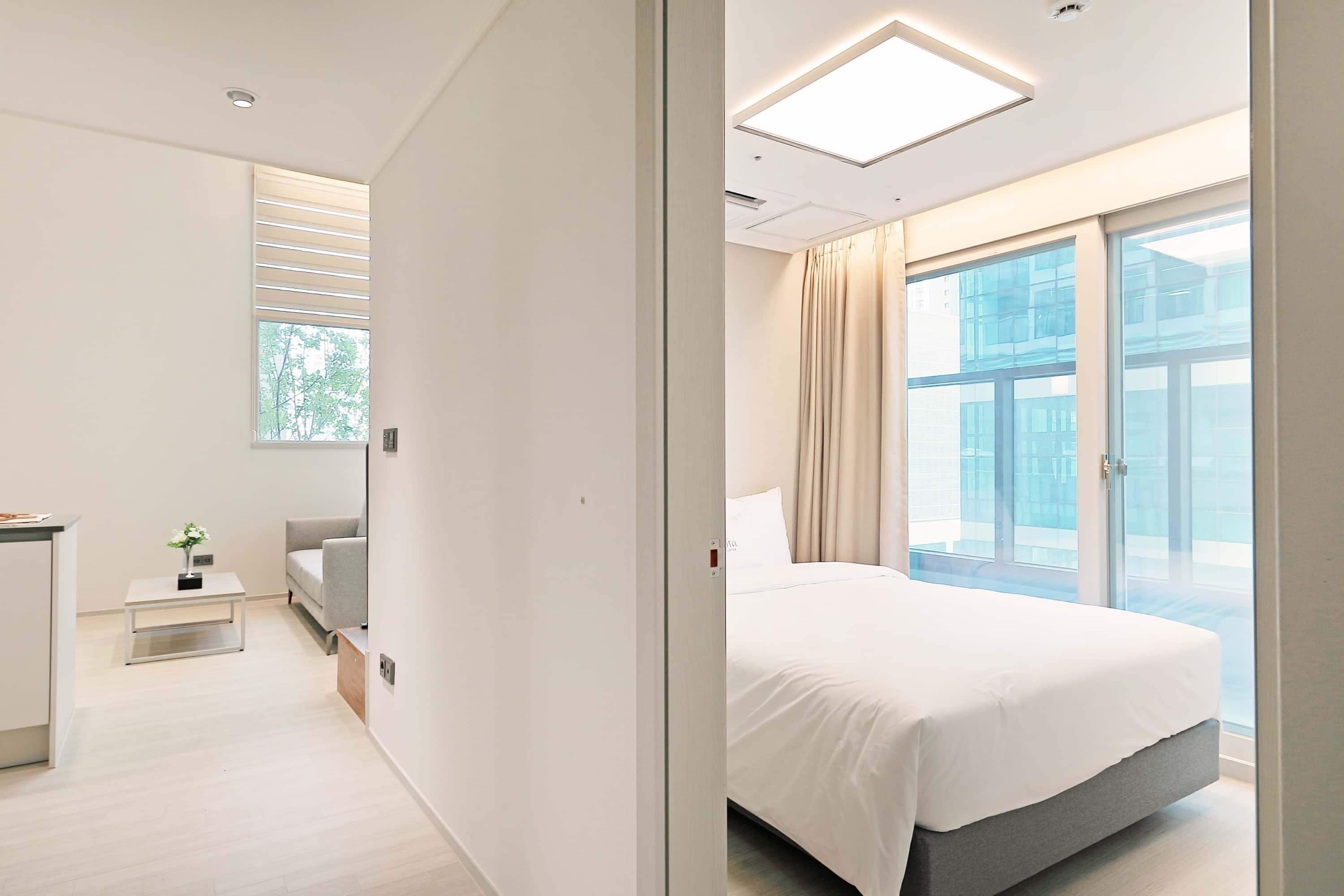A modern hotel room with a bed, a small desk, a sofa, and a large window with a view of the city.