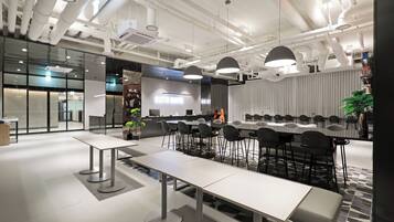 A modern conference room with white tables and chairs, a large screen, and industrial-style ceiling pipes.