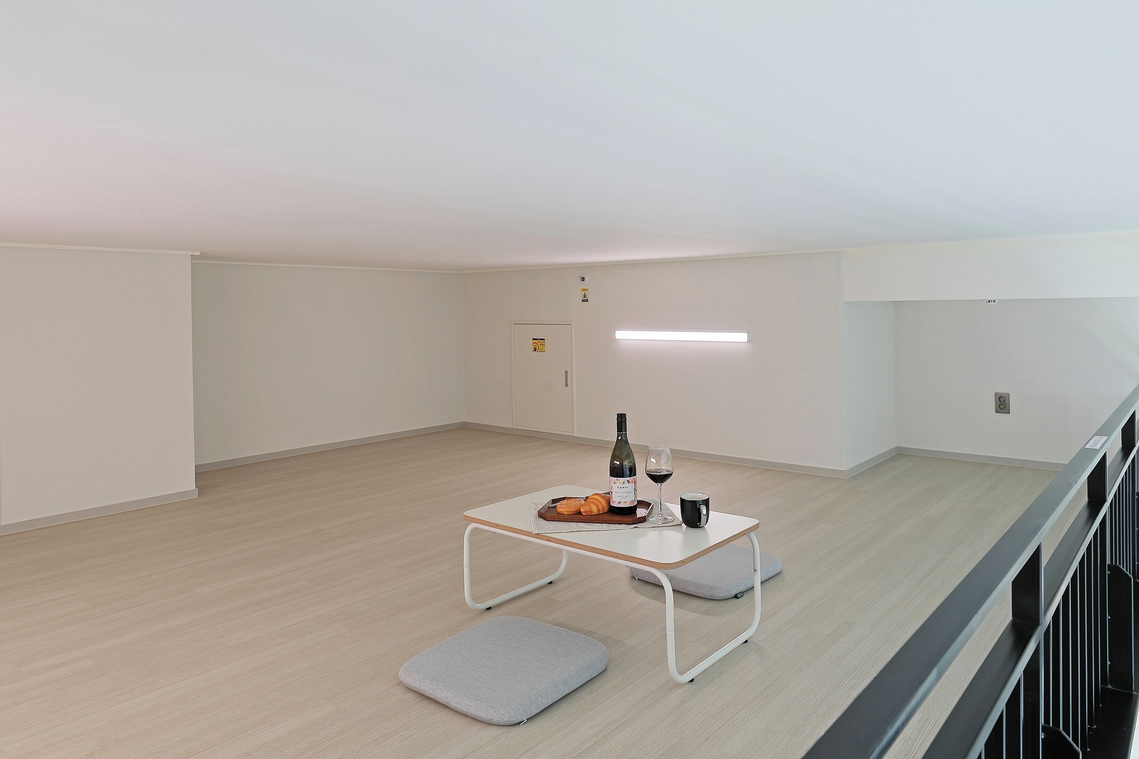 A spacious room with a light wooden floor, a white ceiling, and a small table with a bottle, glass, and bread.