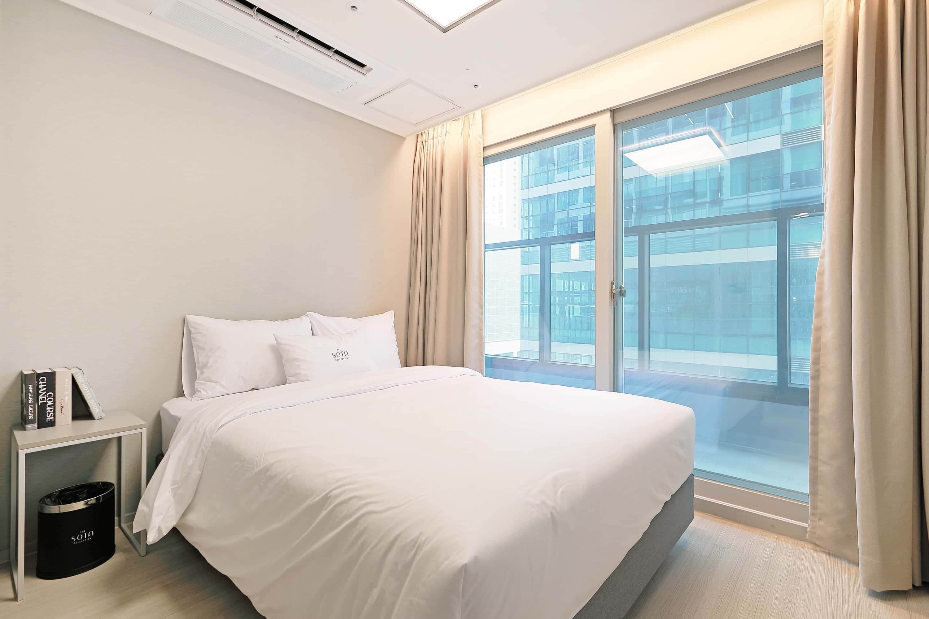 A modern hotel room with a large bed, a bedside table with books, a trash bin, and a large window with curtains.