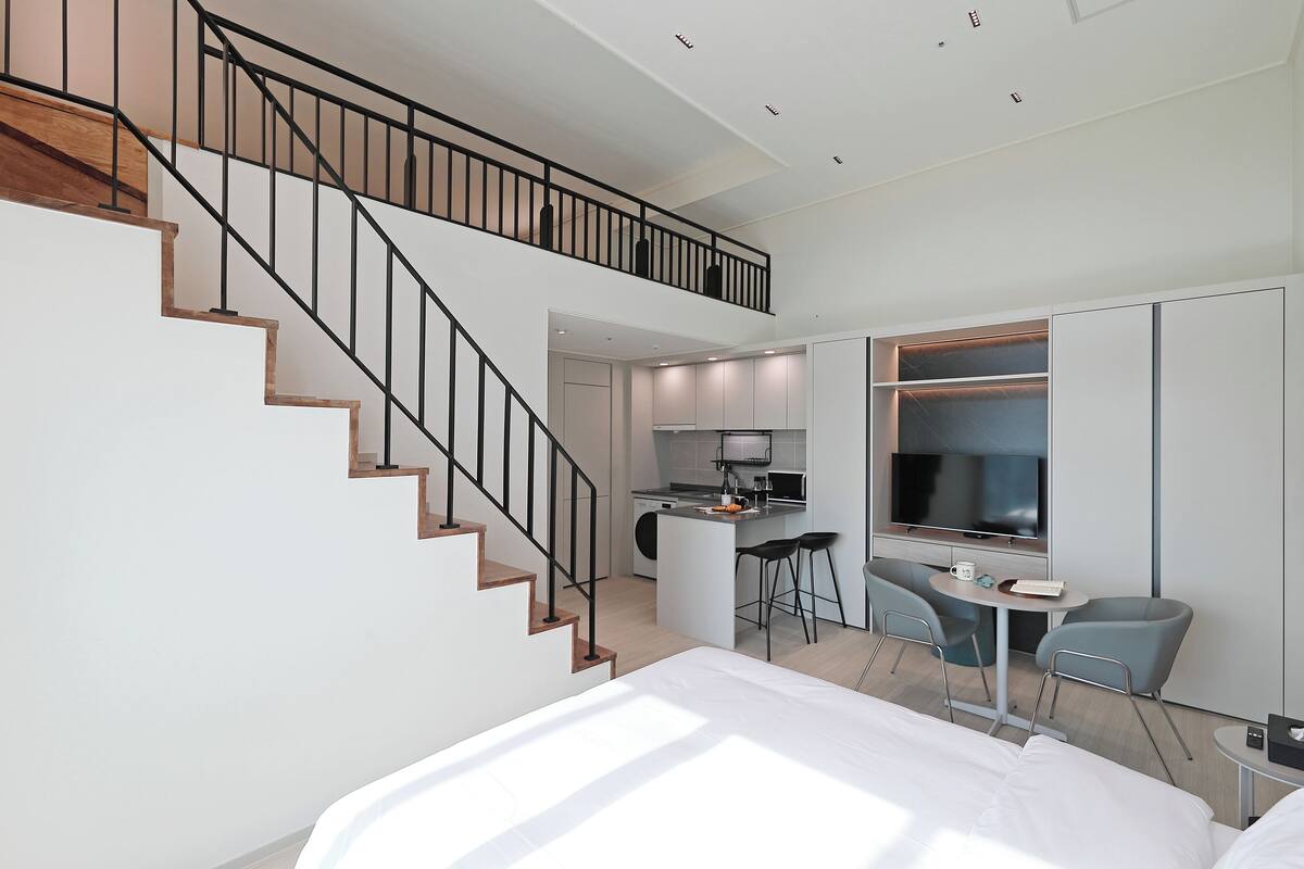 Une chambre d’hôtel moderne dotée d’un escalier, d’un petit coin cuisine, d’une table à manger avec des chaises et d’un grand téléviseur.