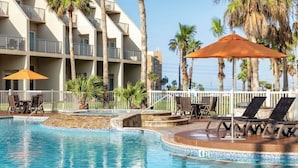 A swimming pool with lounge chairs and umbrellas, surrounded by palm trees and a multi-story building.