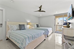 Une chambre avec deux lits, un ventilateur de plafond, un balcon avec vue sur les bâtiments et un bureau avec une chaise.