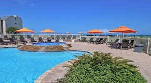 Un espace piscine avec des chaises longues et des parasols, entouré d’une clôture blanche, et un bâtiment en arrière-plan.
