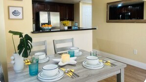 A dining area with a table set for four, a kitchen in the background, and framed artwork on the wall.