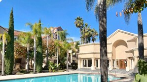 A pool area with a clear blue pool, surrounded by palm trees and a building with a beige facade and arched entrance.