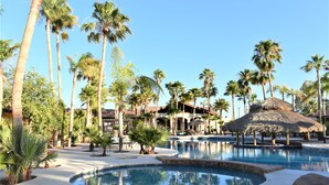 A resort pool area with palm trees, a thatched-roof structure, and a building in the background. - Hacienda Monarcas Resort (Puerto Peñasco)