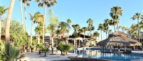 A resort pool area with palm trees, a thatched-roof structure, and a building in the background.