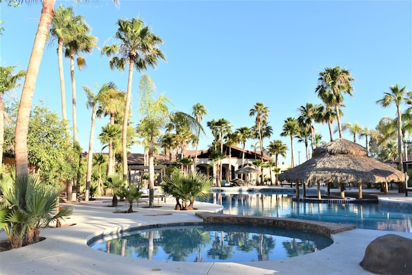 A resort pool area with palm trees, a thatched-roof structure, and a building in the background.