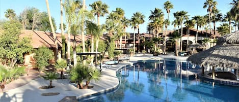 Zona de piscina en un resort con palmeras, una estructura de techo de paja y un cielo azul despejado.