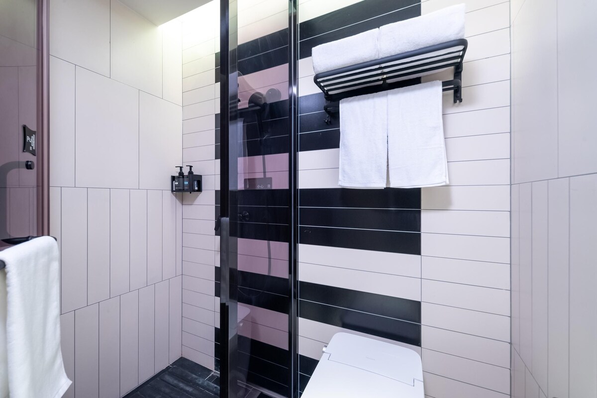 A bathroom with a glass shower enclosure, white and black tiled walls, a white toilet, and a towel rack with a white towel.