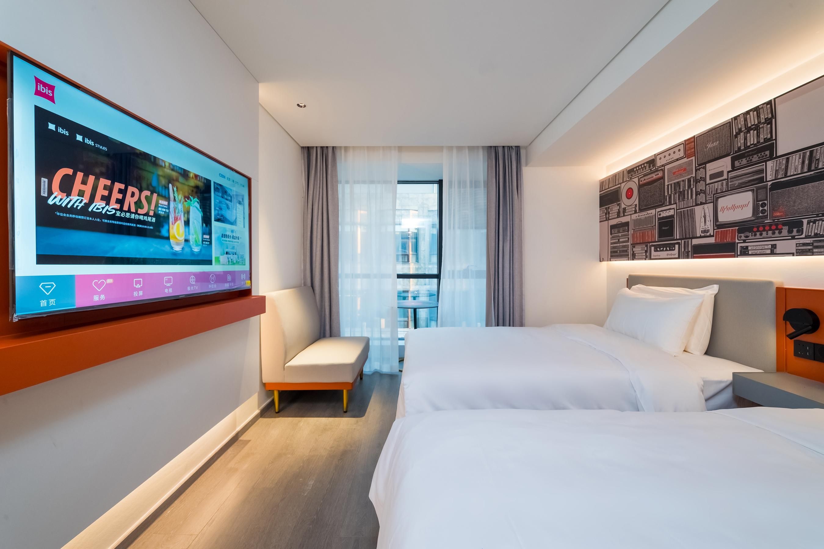 Habitación de hotel con dos camas, un televisor en la pared, un pequeño rincón de descanso y vistas a la ciudad desde la ventana.