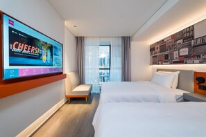 A hotel room with two beds, a TV on the wall, a small seating area, and a view of the city through the window. - Ibis Hotel (Beijing)