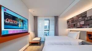 Camera d'albergo con due letti, una TV a parete, una piccola zona salotto e vista sulla città dalla finestra.