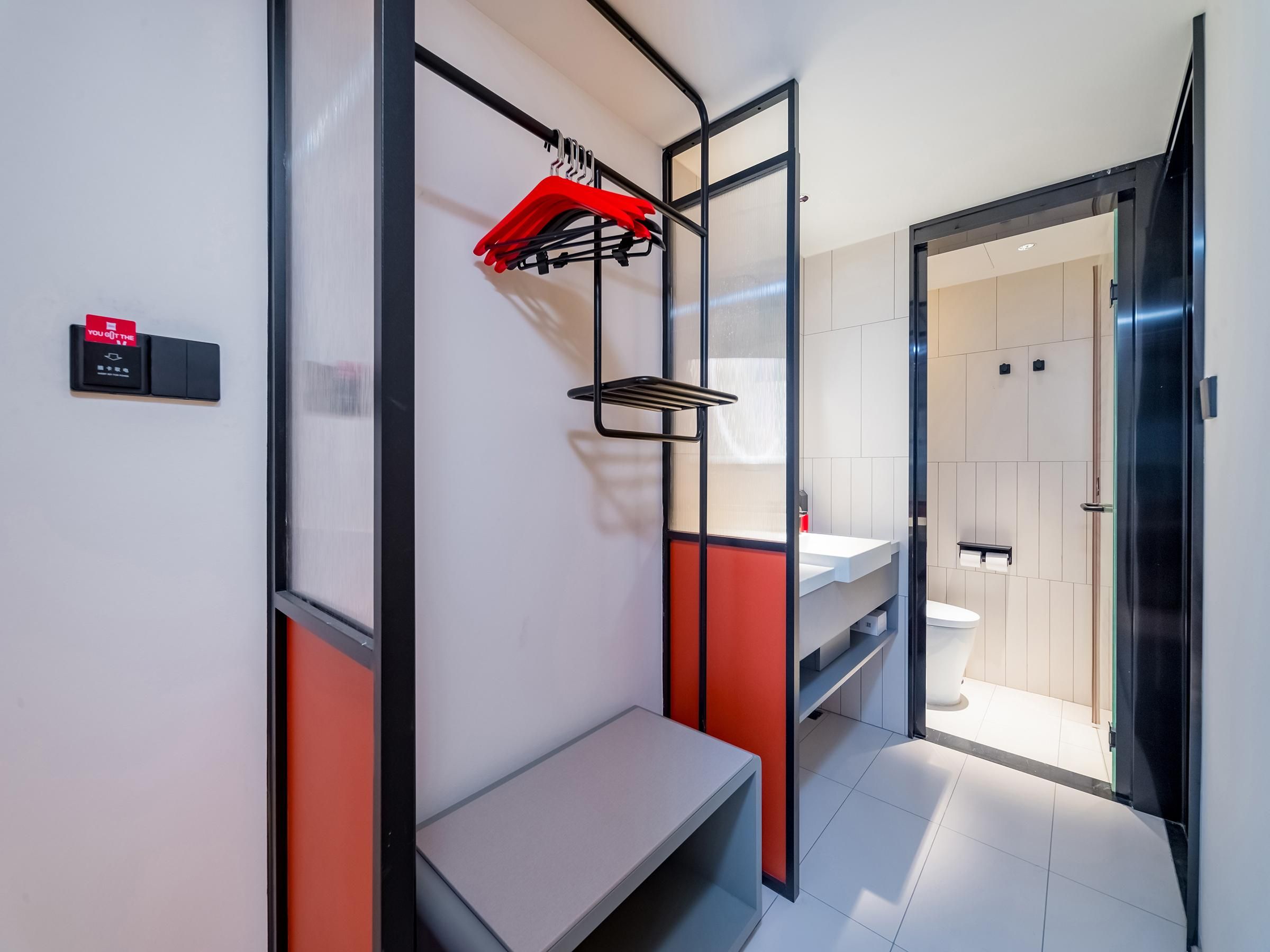 A modern hotel room with a bench, a red hanging rack, and a visible bathroom with a toilet and sink.