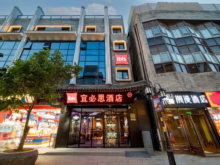 Um hotel com fachada moderna, um letreiro chamativo 'ibis' e um elemento arquitetônico tradicional chinês.