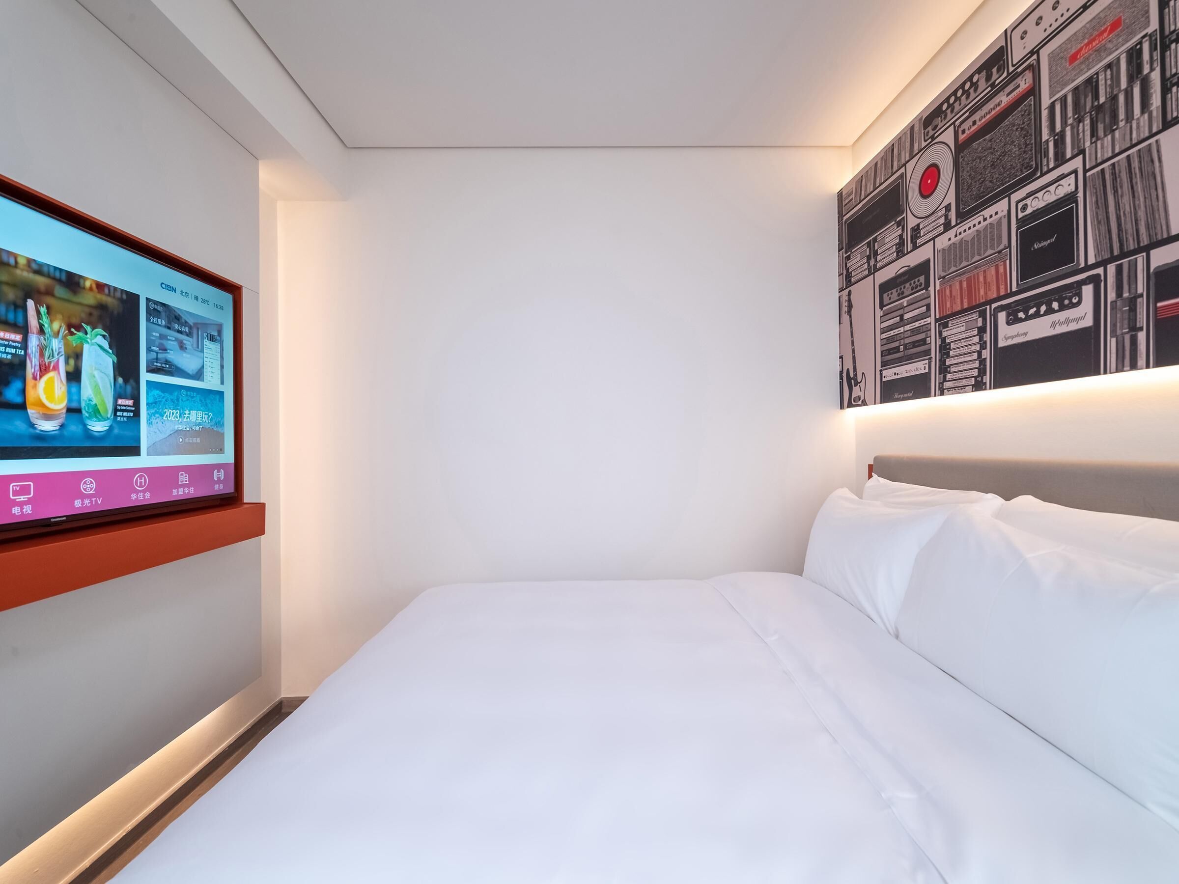 A hotel room with a bed, a mounted TV displaying a menu, and a wall with a pattern of black and white images.