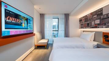 A hotel room with two beds, a TV on the wall, a small seating area, and a view of the city through the window.