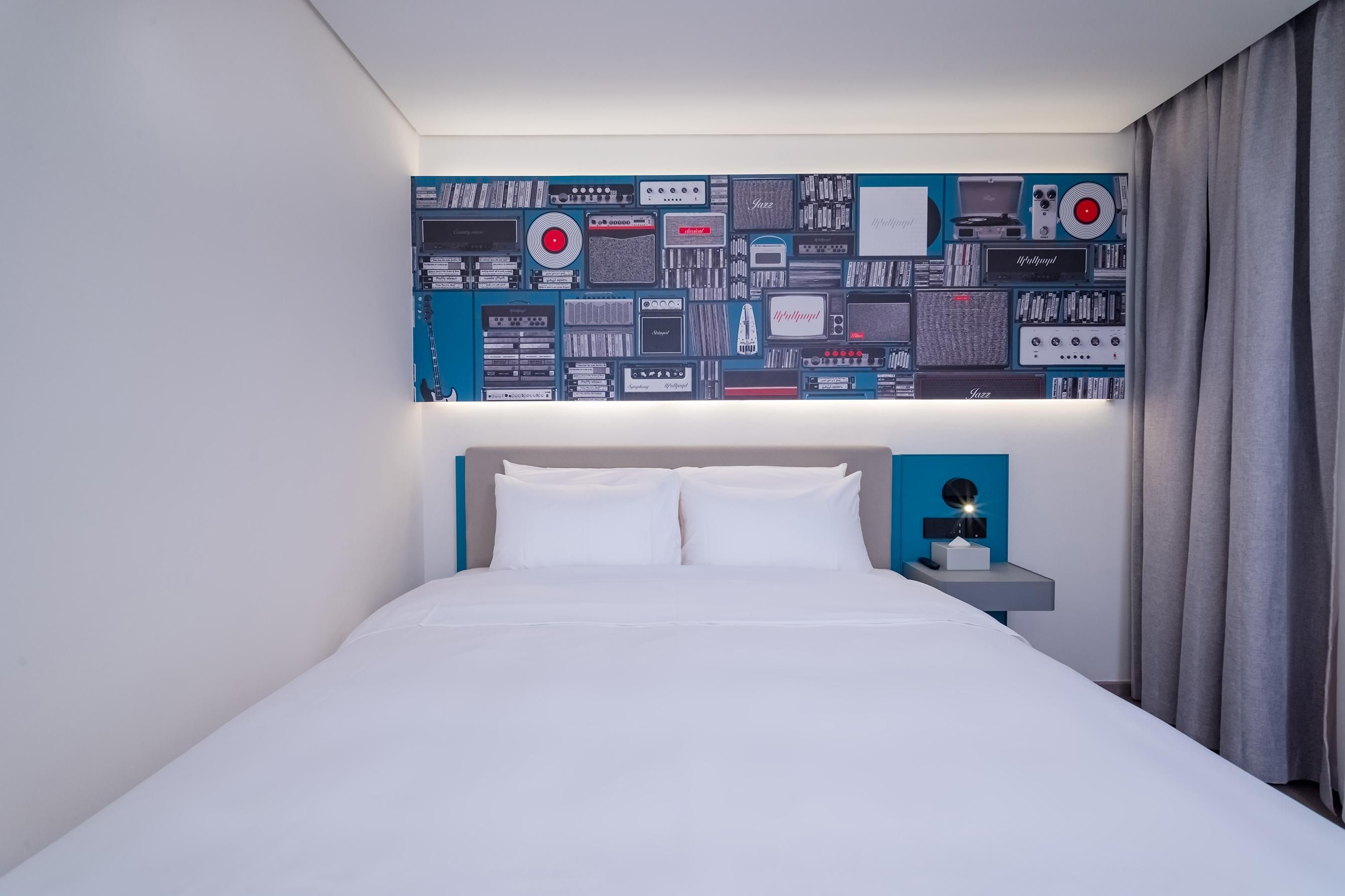A hotel room with a bed, a nightstand with a lamp, and a wall decorated with vintage audio equipment.