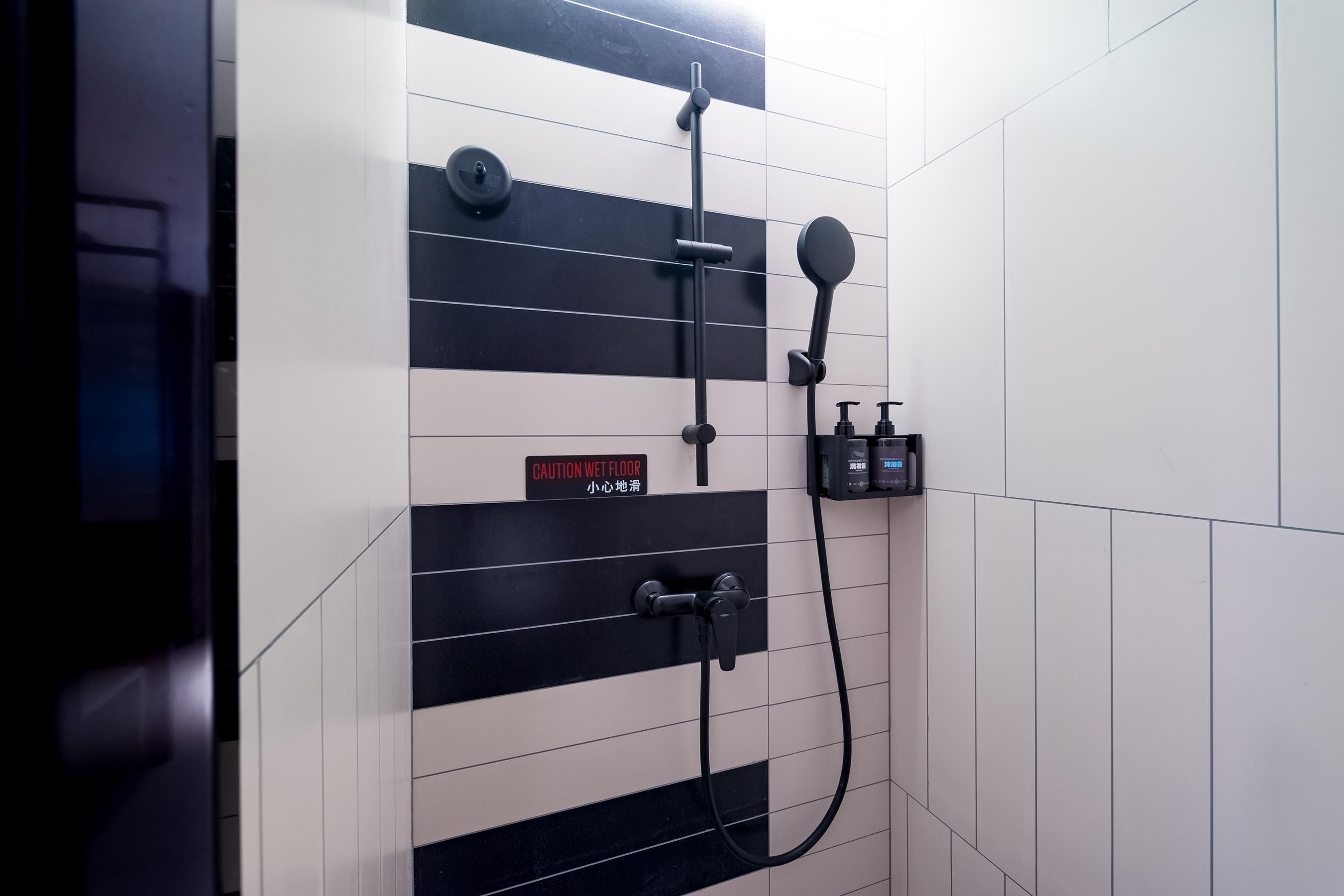 Un baño moderno con paredes de baldosas blanco y negro, un letrero de precaución, un cabezal de ducha manual y un grifo de pared.