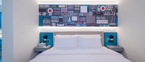 Quarto de hotel moderno com uma cama grande, duas mesas de cabeceira e uma obra de arte na parede com dispositivos eletrônicos.