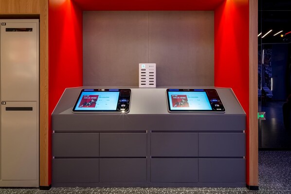 A hotel reception desk with two digital kiosks, a wooden cabinet with a lock, and a red wall. - Ibis Hotel (Beijing)