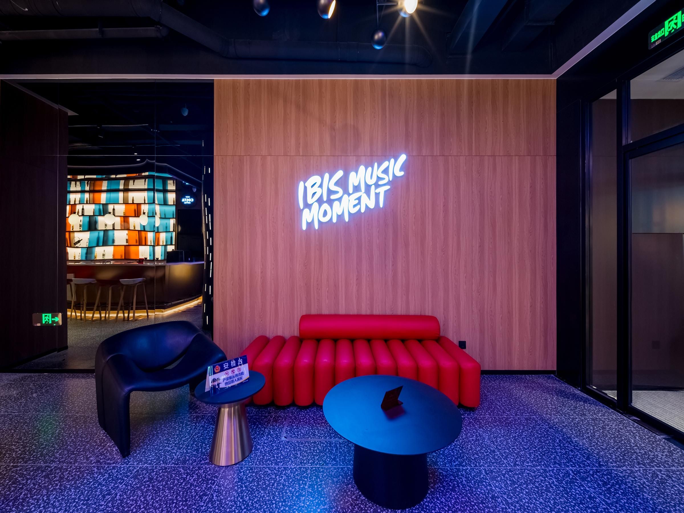 Ein moderner Loungebereich mit einem Neonlichtschild mit der Aufschrift "IBIS MUSIC MOMENT", einem roten Sofa, einem blauen Tisch und einem schwarzen Sessel.