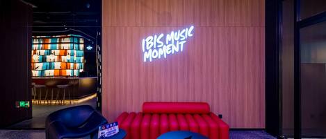 Ein moderner Loungebereich mit einem Neonlichtschild mit der Aufschrift "IBIS MUSIC MOMENT", einem roten Sofa, einem blauen Tisch und einem schwarzen Sessel.