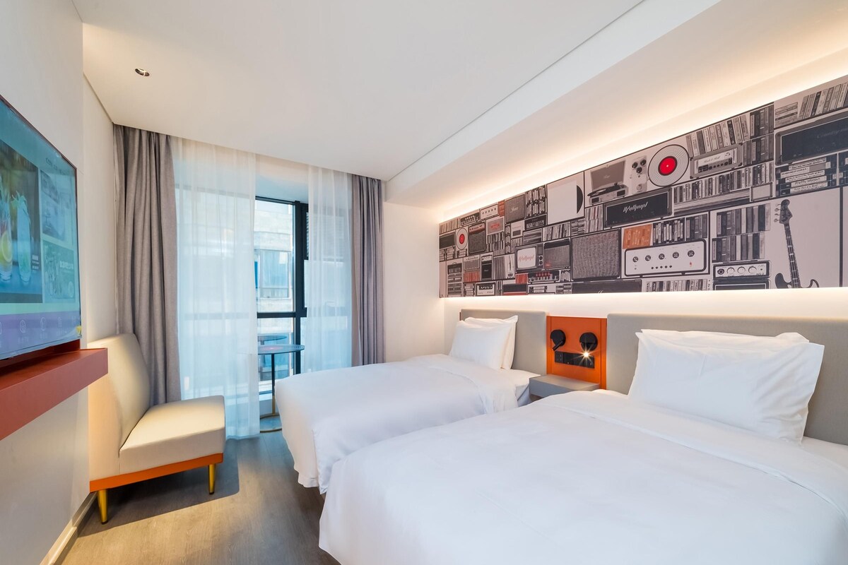 A hotel room with two beds, a TV, a small table, and a large window with curtains.