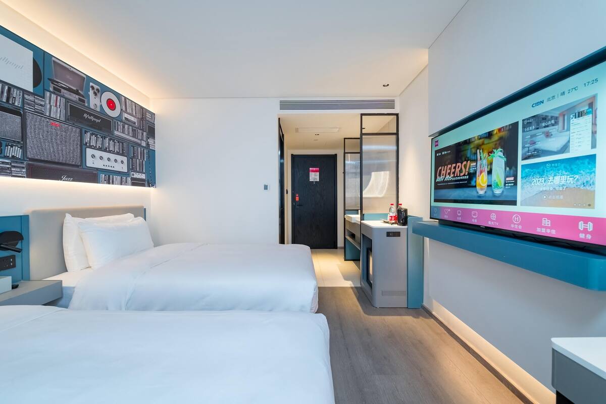Ein Hotelzimmer mit zwei Betten, einem an der Wand montierten Fernseher und einem Schreibtisch mit einem Computer.