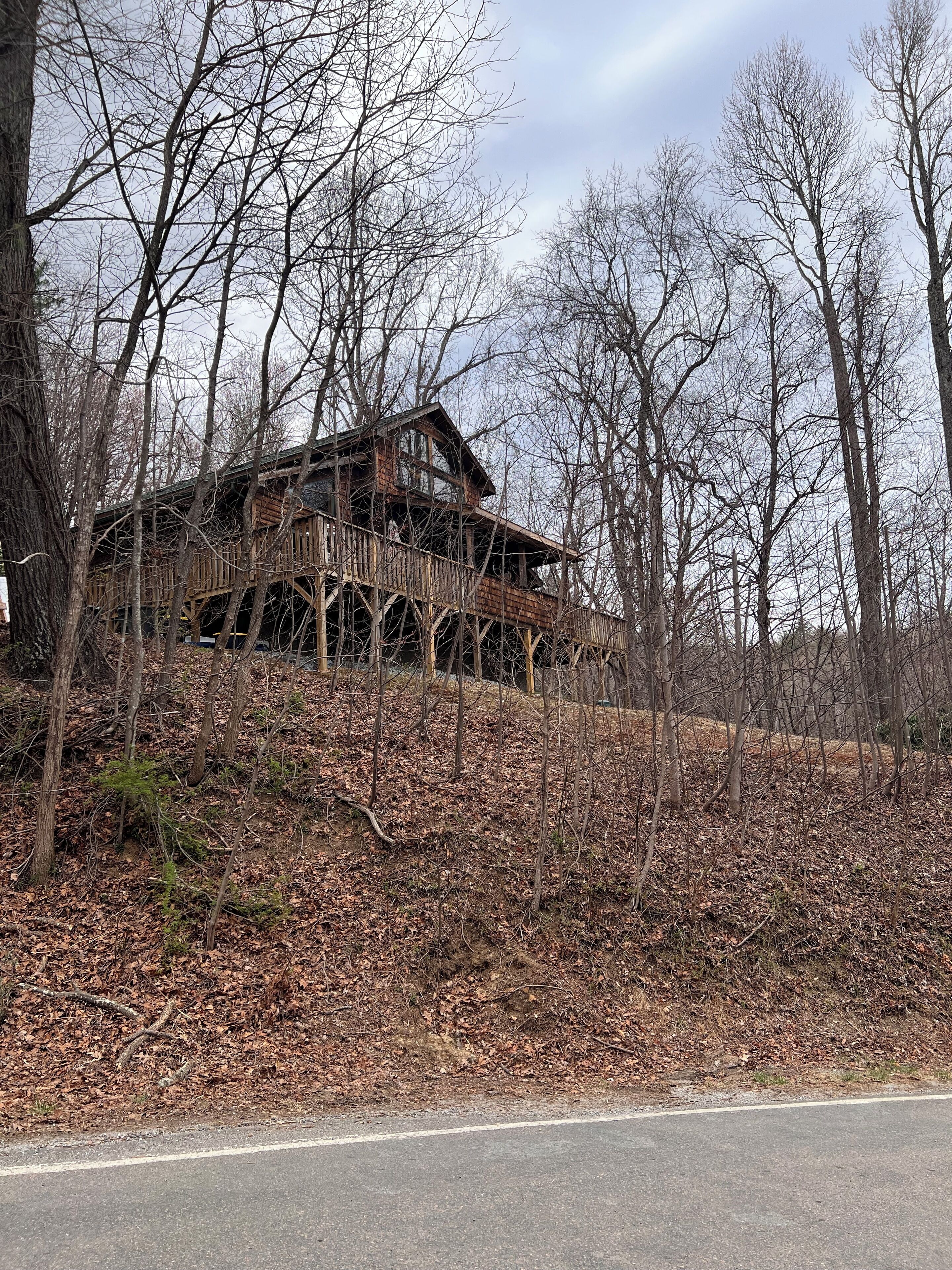 A rustic cabin with a wooden deck, surrounded by leaf-covered ground and bare trees.