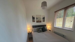 A bedroom with a bed, two bedside lamps, a hanging light fixture, and a window with blinds.