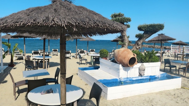 Un área con mesas y sillas al aire libre, una maceta de barro grande y una pequeña fuente de agua en una playa arenosa con sombrillas y palmeras.