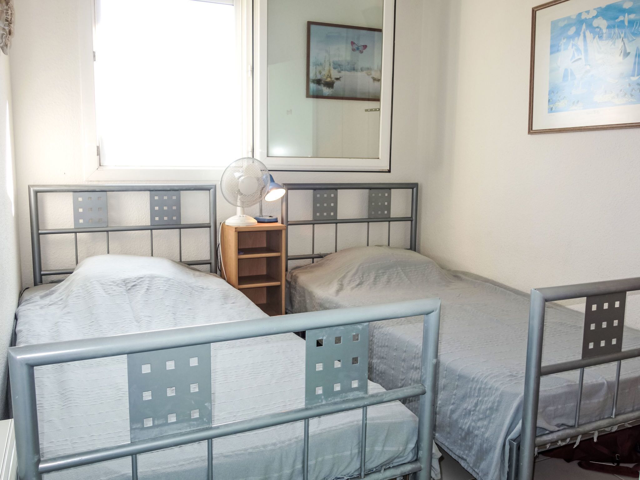 Une chambre double avec deux lits, une table de chevet avec un ventilateur, une fenêtre et un tableau au mur.