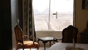 Ein Zimmer mit einem Bett, zwei Stühlen, einem kleinen Tisch und Blick auf Pyramiden durch ein Fenster.