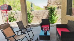 En udendørs terrasse med fletmøbler, en basketballkurv og et lille bord med en blomstervase.