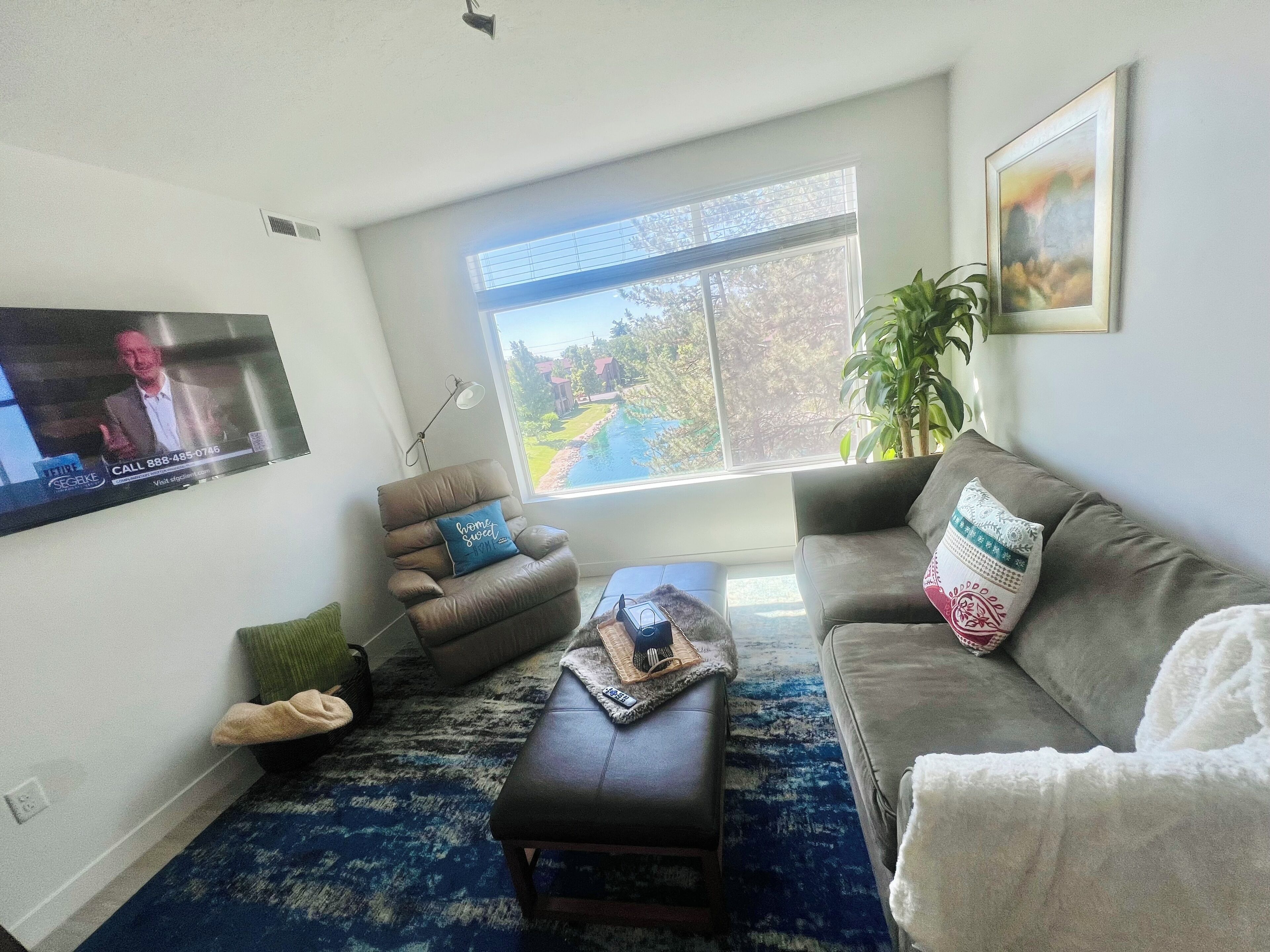 Una sala de estar con un ventanal grande, un televisor de pantalla plana, un sofá gris, un sillón de cuero marrón y un tapete azul estampado.