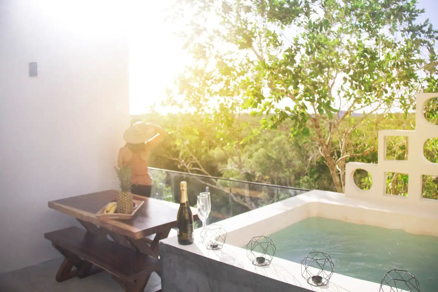 A balcony with a wooden table, a bottle of champagne, and glasses, overlooking a swimming pool and lush greenery.