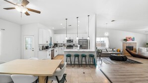 A modern kitchen with white cabinets, a central island with bar stools, a dining area with a wooden table and chairs, and a living area with a sofa and a fireplace. - Estrella Escape, Pool, Spa & Game Room Fun (Desert Hot Springs)