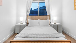 A bedroom with a large bed, two bedside tables, white curtains, and a window with a view of a house. - Estrella Escape, Pool, Spa & Game Room Fun (Desert Hot Springs)