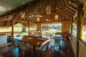 Een overdekt buiten eetgedeelte met houten tafels en stoelen, een rieten dak en uitzicht op het omliggende landschap.