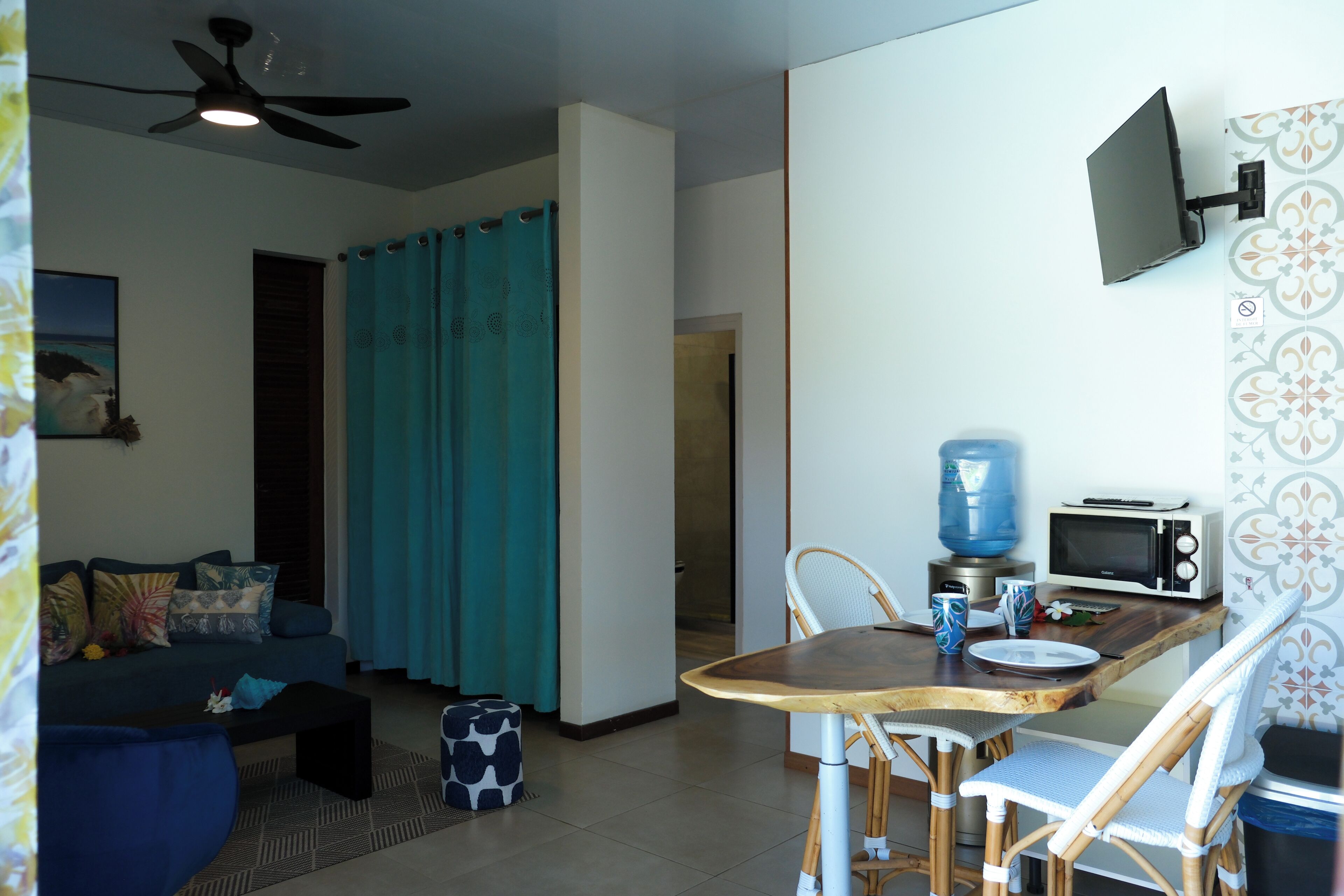 Una sala de estar con sofá, una mesa de centro, una mesa de comedor con sillas, un ventilador de techo, un televisor montado en la pared y una zona de cocina con microondas.
