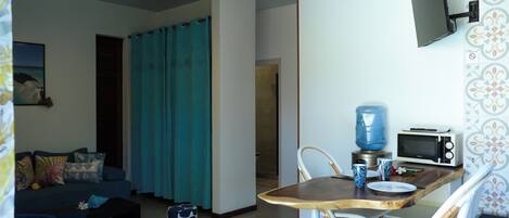 Una sala de estar con sofá, una mesa de centro, una mesa de comedor con sillas, un ventilador de techo, un televisor montado en la pared y una zona de cocina con microondas.