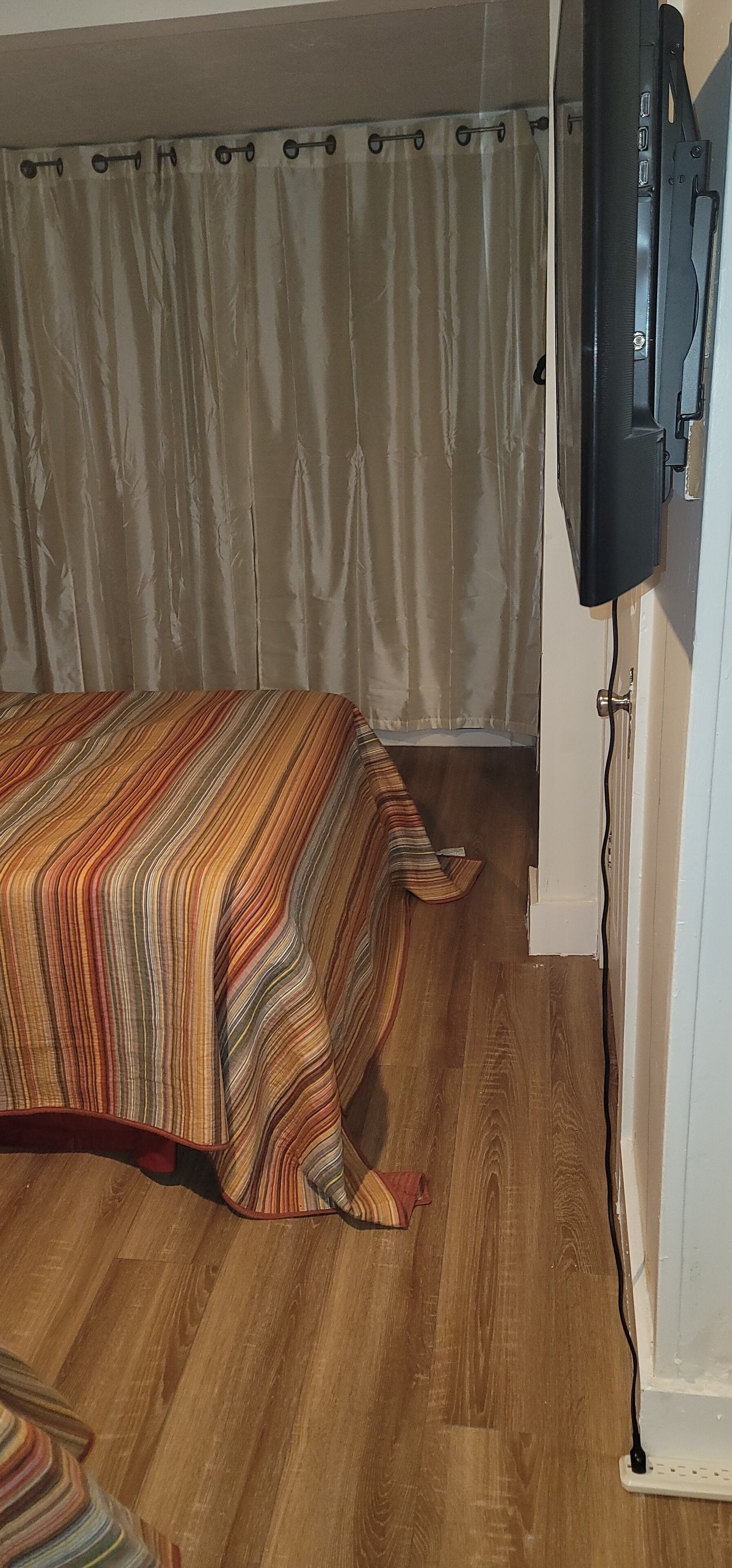 Un dormitorio con una cama, suelo de madera, una cortina y un televisor montado en la pared.