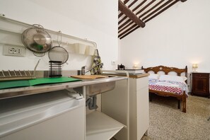 A kitchen with a sink, a cutting board, and various kitchen utensils. There is a bed with a wooden headboard and a nightstand with a lamp.