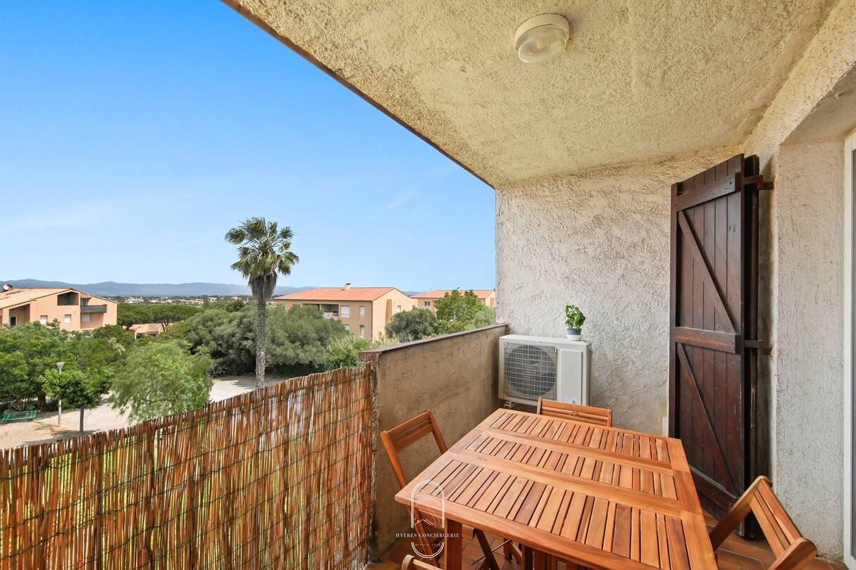 A balcony with a wooden table and chairs, an air conditioning unit, and a view of buildings and trees.