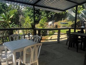 Eine Terrasse im Freien mit Tischen und Stühlen, umgeben von üppiger Bepflanzung und einem Metalldach.