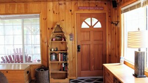 A cabin interior with a wooden door, a bookshelf, a window with a view of outdoor chairs, and a lamp on a wooden table. - Sugar Bear (Big Bear City)