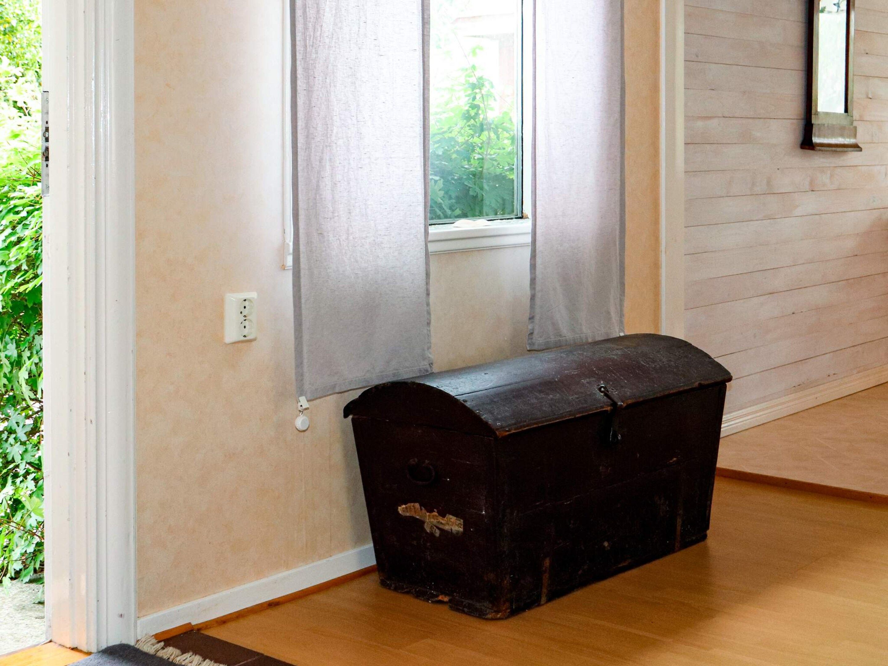 Una habitación con suelo de madera, una ventana con cortinas y un cofre de madera oscura.